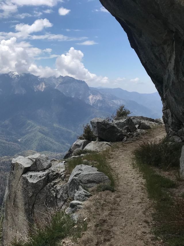 A narrow, winding dirt trail along a steep cliff edge with expansive views of rugged mountains and valleys.