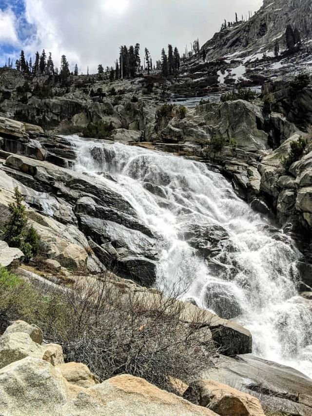 A powerful, multi-tiered waterfall cascading over grey, rocky slopes surrounded by scattered pines and snow patches.